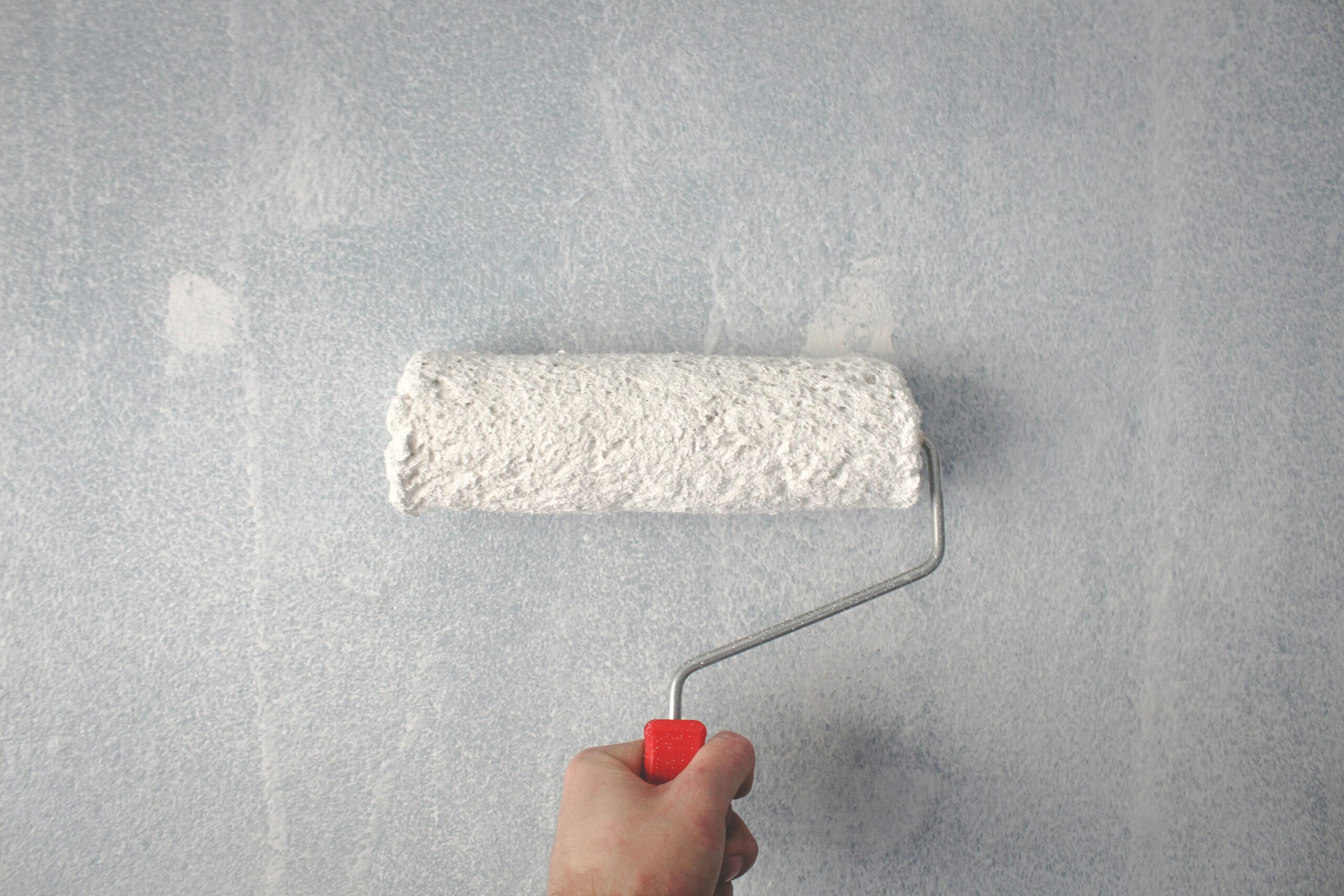 Home A person's hand using a paint roller to apply white paint on a wall.